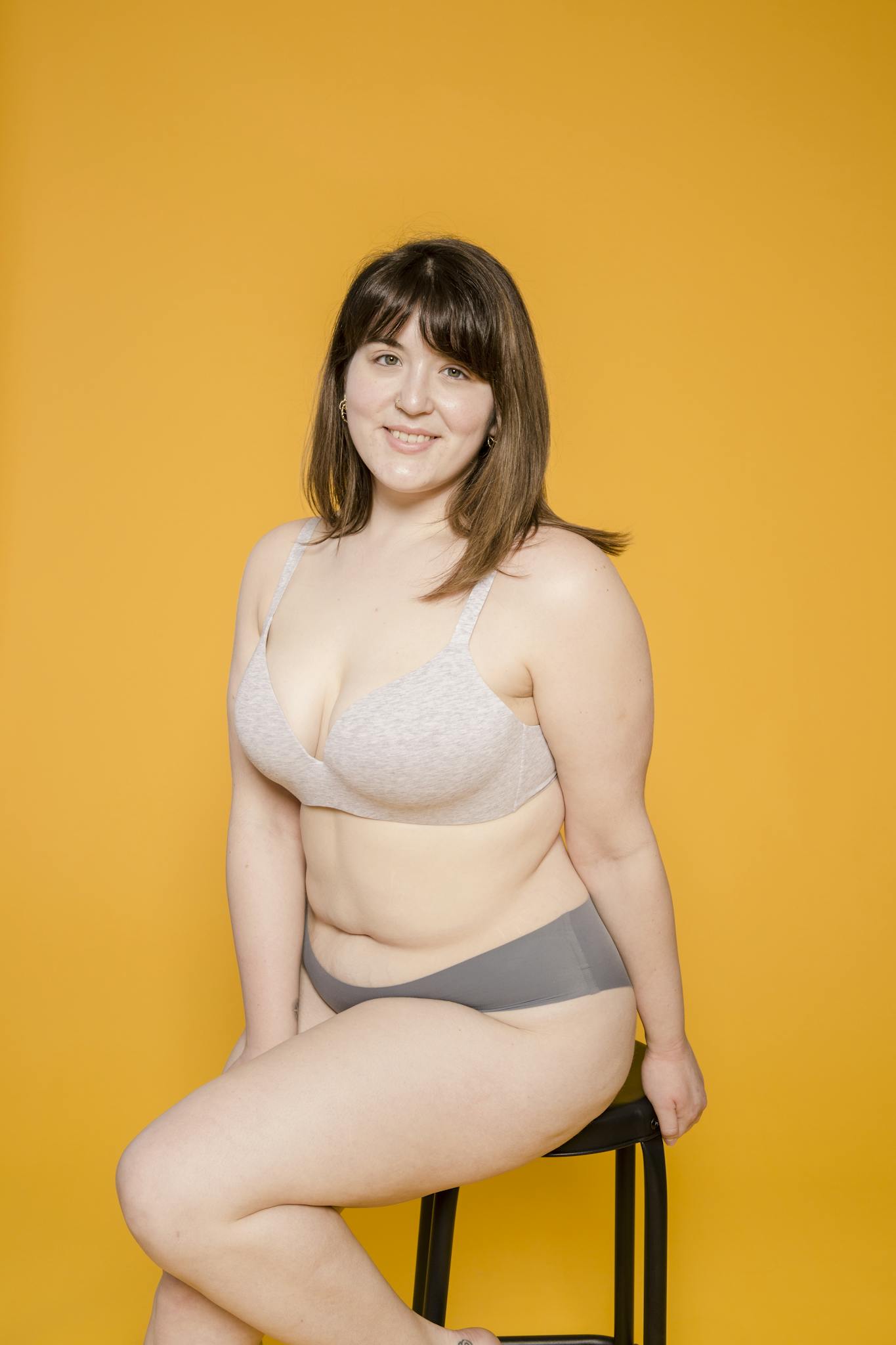 Portrait of a smiling plus size woman seated on a stool in lingerie, embracing body positivity against a bright yellow backdrop.