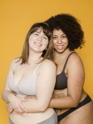 Two diverse women in underwear, smiling and embracing against a vibrant yellow background.
