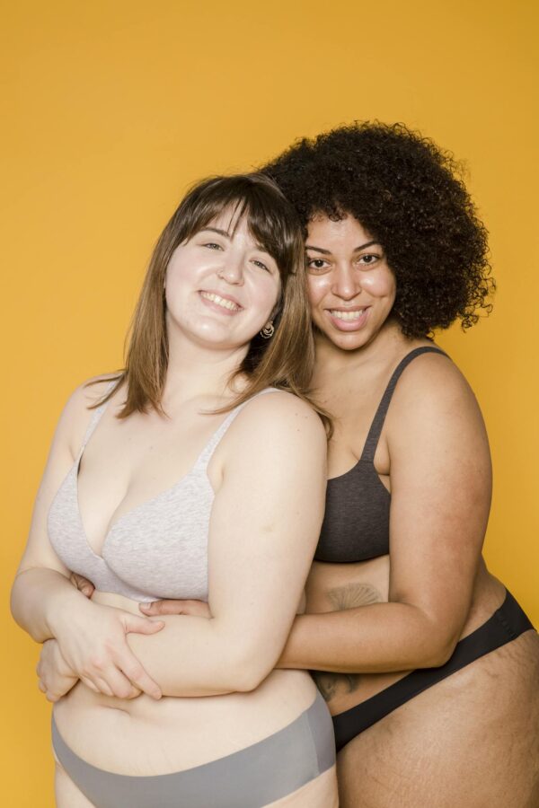 Two diverse women in underwear, smiling and embracing against a vibrant yellow background.